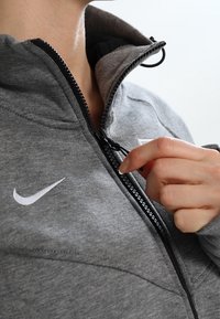 Chaqueta gris con cremallera y cuello alto, con una cremallera negra y un logo blanco de Nike en el pecho. Textura de tejido suave.