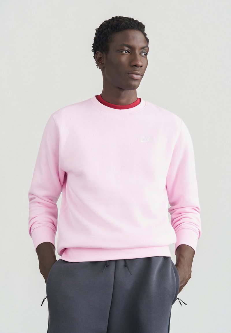 Sweatshirt cor de rosa claro com decote redondo e punhos canelados, com um pequeno logótipo branco da Nike no peito, combinado com calças cinza escuro.