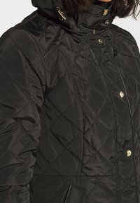 Svart quiltad jacka med hög krage, utsmyckad med guldfärgade detaljer och en dragkedjeficka. Tillverkad av ett glansigt och slätt material.