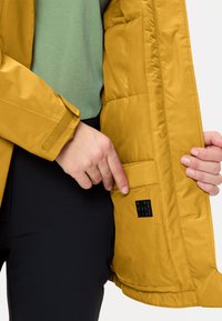 Giacca imbottita gialla con un esterno liscio, dotata di un'etichetta nera per il riciclo in una tasca interna. La persona indossa una maglietta verde sotto.