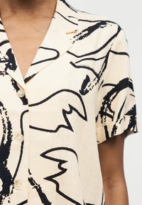 Chemise beige clair à manches courtes avec des motifs abstraits en lignes noires et des boutons sur le devant, portée par une personne à la peau marron moyen.