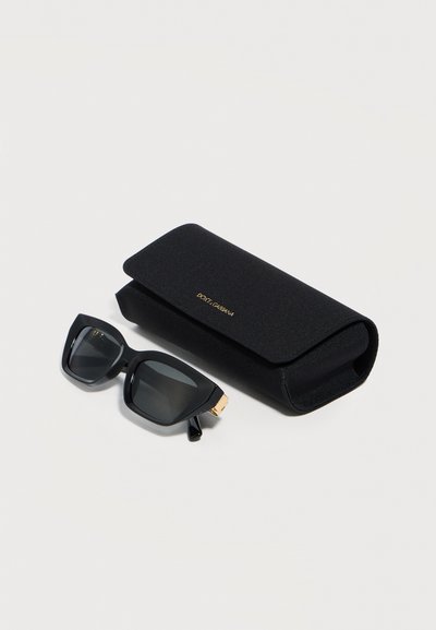 Μαύρα γυαλιά ηλίου με μαύρους φακούς σε σχήμα γατίσιου ματιού δίπλα σε μαύρη θήκη Dolce & Gabbana σε λευκό φόντο.