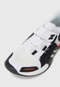 Sneaker bianco in rete con accenti neri e suola in gomma. Presenta un sistema di allacciatura sicuro e il logo "new balance" in arancione. Design testurizzato.