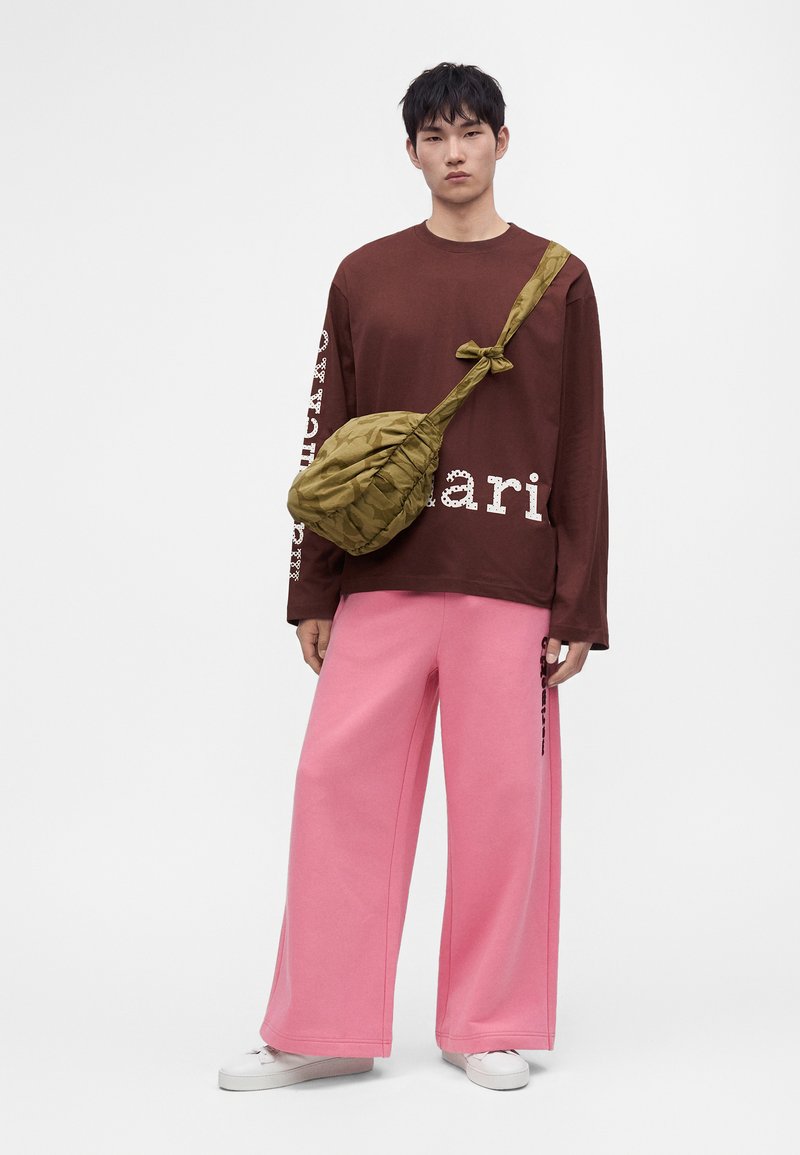Νέος άντρας στέκεται φορώντας καφέ oversized πουκάμισο με μακριά μανίκια, ροζ παντελόνια με φαρδιά πόδια, λευκά παπούτσια και πράσινη τσάντα ώμου με λεπτομέρεια κόμπο.