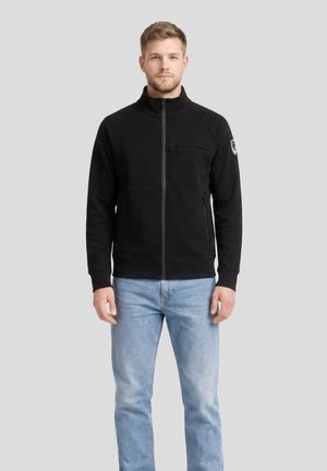 Man draagt een zwart jasje met rits, met borst- en zijzakken, lichtblauwe spijkerbroek, staand tegen een lichtgrijze achtergrond.