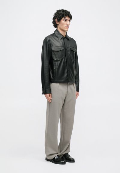 Μαύρο δερμάτινο jacket με δύο μπροστινές τσέπες, συνδυασμένο με ανοιχτό γκρι πλισέ παντελόνι και μαύρες loafers. Λείο υφή και ραμμένη εφαρμογή.