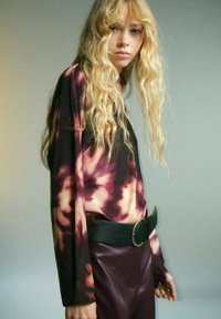 Blusa de manga larga con un patrón de tie-dye en púrpura y crema, combinada con pantalones de cuero sintético burdeos de cintura alta y un cinturón negro ancho.