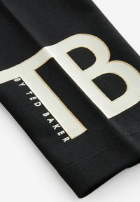 Melns audums ar izteiktu balto un zelta logotipu. Burti ir treknrakstā, ar gludām tekstūrām un kontrastējošām krāsām.