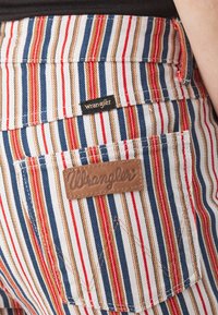 Pantalon rayé en blanc, bleu, rouge et beige avec des étiquettes visibles de marque Wrangler en cuir et en tissu sur la poche arrière.