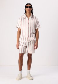 Kortærmet skjorte og shorts sæt i struktureret kremfarvet stof med røde lodrette striber. Fuldført med neutrale sneakers og solbriller.