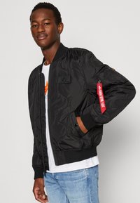 Mustat tikattu bomber-takki, jossa on ribbireunat, vetoketjusuljenta ja punainen kangaslabel vasemmassa hihassa. Paita on valkoinen t-paita.