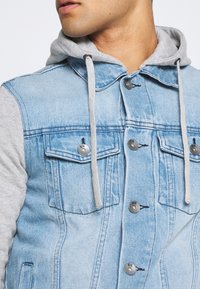 Chaqueta de mezclilla con capucha gris. Mezclilla azul clara con bolsillos frontales y detalles de botones plateados. Capucha suave gris con cordones.