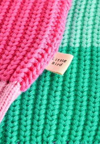 Strickstoff in Pink und Grün mit einem strukturierten Muster. Ein Etikett mit der Aufschrift "Little Bird" ist teilweise im grünen Abschnitt sichtbar.