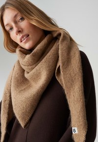 Brauner Wollschal mit dreieckiger Form, weicher Textur und einem kleinen Etikett in der Ecke. Locker um den Hals getragen.