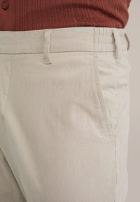 Pantalones de pana beige con finas rayas verticales, que presentan un frente plano, bolsillos laterales y una cintura con pasadores para cinturón.