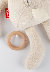 Stofftier aus weichem, beigem Material mit strukturierter Oberfläche; verfügt über einen Holzring, der mit einem weißen Band befestigt ist, sowie einem Stoffetikett.