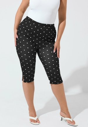 Femme portant un pantalon capri à pois noirs et un top blanc sans manches, debout les mains sur les hanches, sandales blanches à talons visibles.