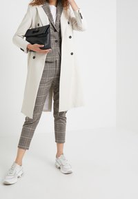 Kobieta w beżowym długim płaszczu, w kratkowym garniturze, białych trampkach, trzymająca czarną kopertówkę na tle białej ściany.