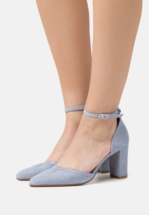 Zapatos de ante azul claro con tacón cuadrado, correa en el tobillo y punta afilada, que presentan un diseño decorativo de costura en la zona de la punta.