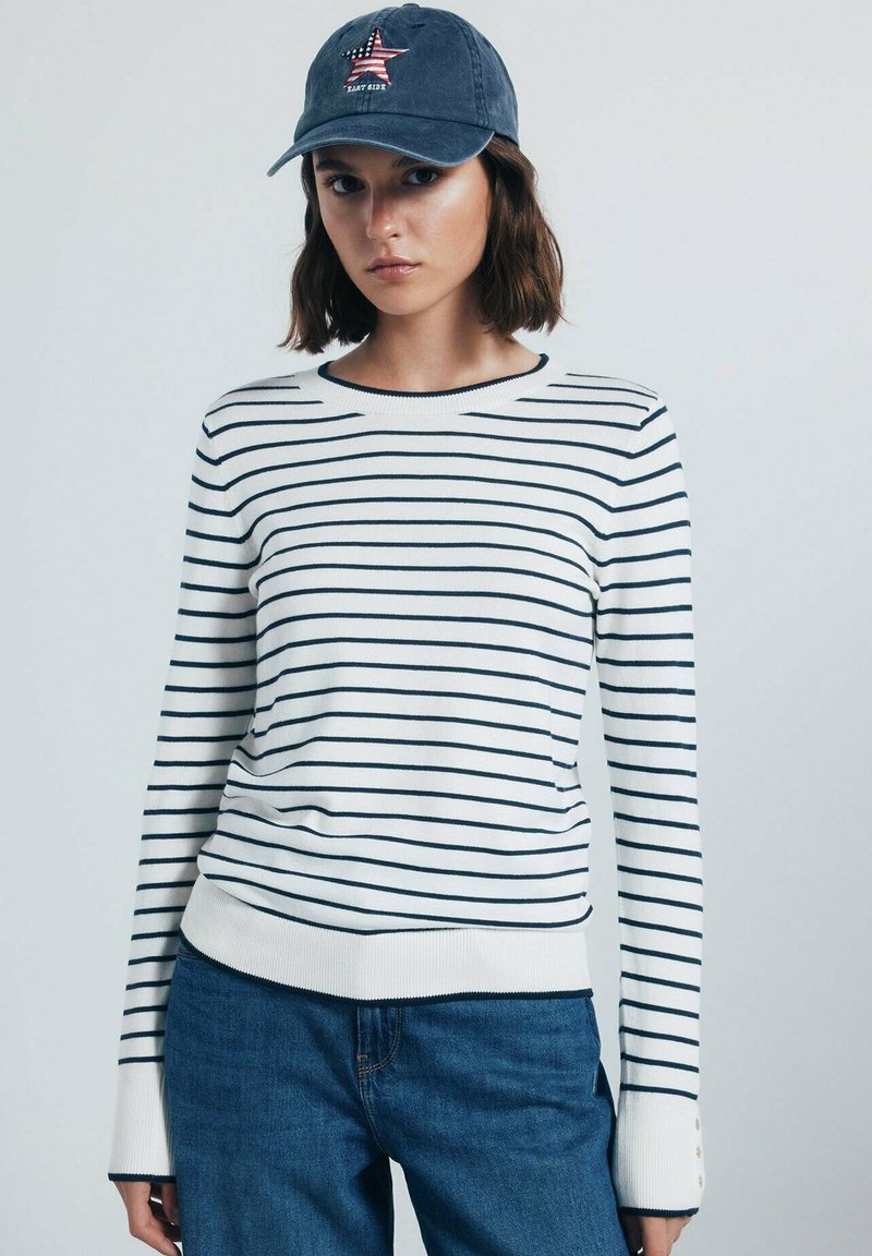 Maglione a righe bianche e navy con maniche lunghe, polsini e orlo a coste. Abbinato a jeans blu e a un cappellino blu con un design a stella.