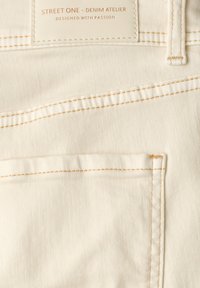 Tissu en denim couleur crème avec des surpiqûres couleur fauve, montrant une poche arrière et une étiquette indiquant « STREET ONE - DENIM ATELIER DESIGNED WITH PASSION. »