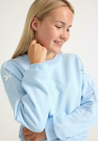 Felpa azzurra realizzata in tessuto morbido, con piping bianco lungo le cuciture e dettagli decorativi a forma di fiocco sulle spalle.