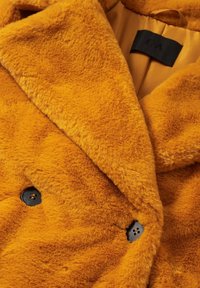 Manteau orange doux et en peluche avec de grands revers et des boutons noirs, montrant une doublure intérieure partielle et une étiquette de vêtement noire.