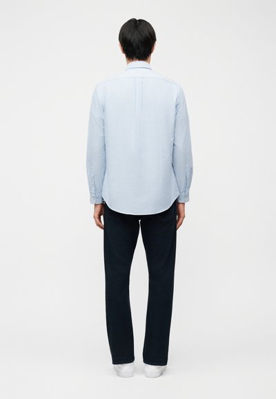 Άνδρας με την πλάτη γυρισμένη, φοράει ανοιχτό μπλε ριγέ μακρυμάνικο πουκάμισο, σκουρόχρωμο παντελόνι και λευκά παπούτσια, μπροστά σε απλό λευκό φόντο.