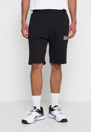Mann trägt schwarze knielange Shorts mit weißem Text am linken Bein, weiße Socken und weiße-schwarze Sportschuhe und steht auf einem grauen Boden.