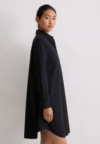 Kobieta w luźnej czarnej sukience z długim rękawem stoi bokiem do gładkiej białej ściany, patrząc w obiektyw.