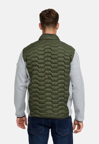 Mand iført grøn quiltet vest over grå langærmet skjorte, set bagfra, stående mod en ensartet hvid baggrund.