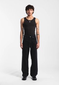 Musta ribbattu toppi, rentoistuiset mustat farkut, hopeinen kaulakoru suorakulmaisella riipuksella, seisoo yksinkertaisella taustalla.