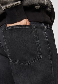 Black denim jeans with a back pocket detail. The fabric has a slight texture, with visible stitching and a slim fit design.