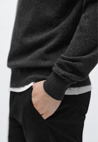 Tmavě šedý svetr s žebrovanými manžetami a texturou těla, vrstvený přes bílou košili, kombinovaný s černými kalhotami, ruka v kapse.