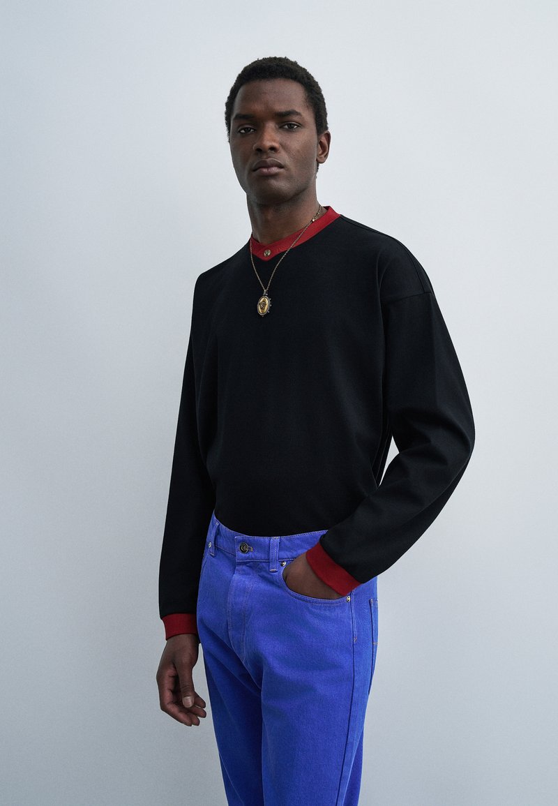 Homme portant une chemise noire à manches longues avec col et poignets rouges, pantalon bleu et un collier pendentif, debout sur un fond gris clair.
