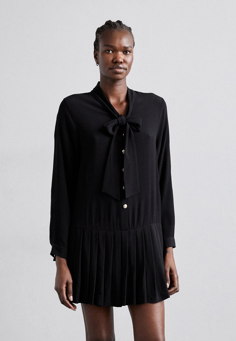 Robe noire à manches longues, jupe plissée et col cravate. Présente des boutons dorés et une texture lisse. Design élégant et minimal.