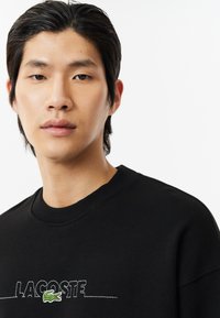 Sudadera negra de cuello redondo, con el texto bordado "LACOSTE" y un logotipo de cocodrilo en verde. Tejido suave, ajuste relajado.