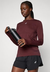 Mujer con camisa de manga larga burdeos y pantalones cortos negros sosteniendo y abriendo una botella de agua de acero inoxidable negra frente a un fondo neutro.