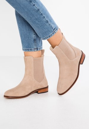 Bottines en daim beige avec panneaux latéraux élastiques et semelles en bois, portées avec un jean bleu retroussé sur un sol blanc.