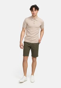 Camiseta de polo de punto beige con tres botones, combinada con pantalones cortos verde oliva y zapatillas blancas, creando un atuendo casual.