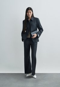 Räätälöity tumma liituraitapuku, jossa on leveät liehuttavat housut ja kauluksellinen takki. Yhdistetty beigeen toppiin ja mustaan clutch-laukkuun. Valkoiset terävät korot.