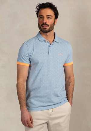 Man met donker haar en baard, draagt een lichtblauw patroon polo met oranje mouwboorden en beige broek tegen een effen achtergrond.