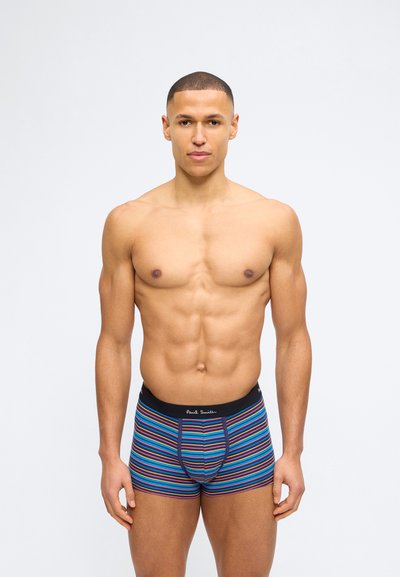 Μυώδης άνδρας όρθιος, φορώντας πολύχρωμα ριγέ μποξεράκια με μαύρη μέση, μπροστά σε απλό φωτεινό φόντο.