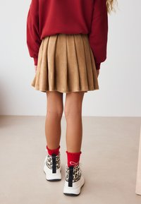 Jupe plissée en velours côtelé couleur bronze associée à des baskets blanches à imprimé léopard. Chaussettes rouges à volants visibles au-dessus des chaussures.