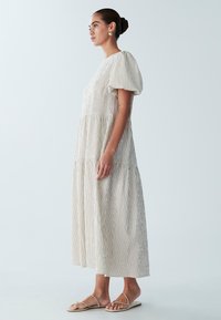 Robe maxi à manches bouffantes, avec des rayures verticales beige et blanches, tissu texturé et silhouette fluide. Portée avec des sandales plates.