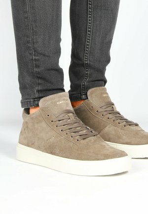 Zapatillas altas de ante beige con cordones planos, un acabado texturizado y una suela de goma blanca, combinadas con jeans oscuros ajustados.