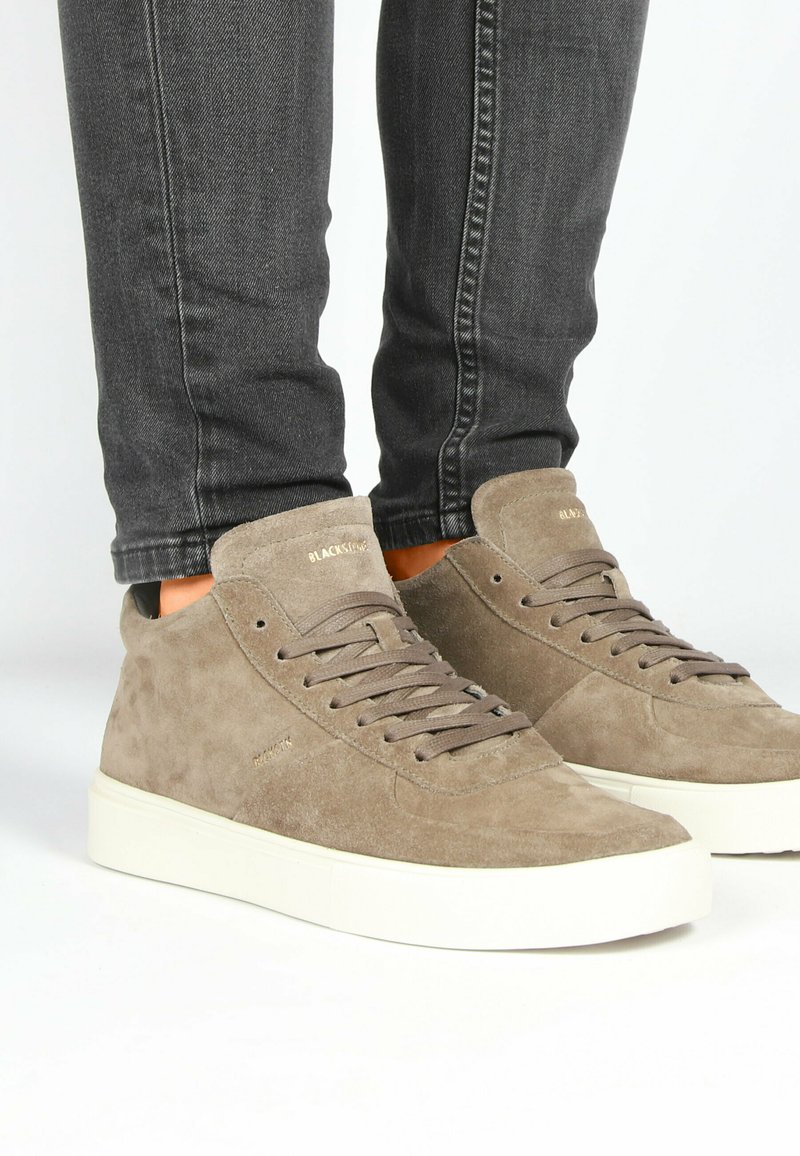 Baskets montantes en daim beige avec des lacets plats, une finition texturée et une semelle en caoutchouc blanche, associées à un jean slim noir.