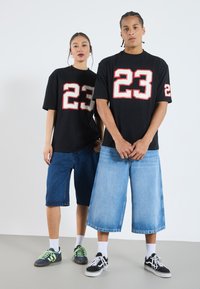 Svarta t-shirts med fet röd och vit siffra "23," kombinerat med lösa denimshorts. Sneakers med gröna accenter och vita strumpor.