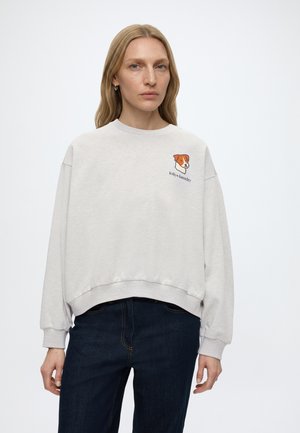 Frau mit glatten blonden Haaren, die einen hellgrauen Pullover mit einer kleinen braun-weißen Hundegrafik und Text auf der Brust trägt, kombiniert mit dunklen Jeans.