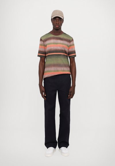 Short-sleeve, ribbed knit t-shirt in green, orange, and brown stripes, paired with navy pants and white sneakers, topped with a beige cap.